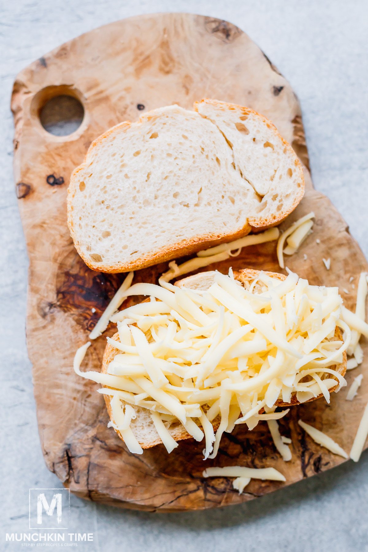 Two slices of bread on a cutting board, one slice has grated mozzarella cheese.